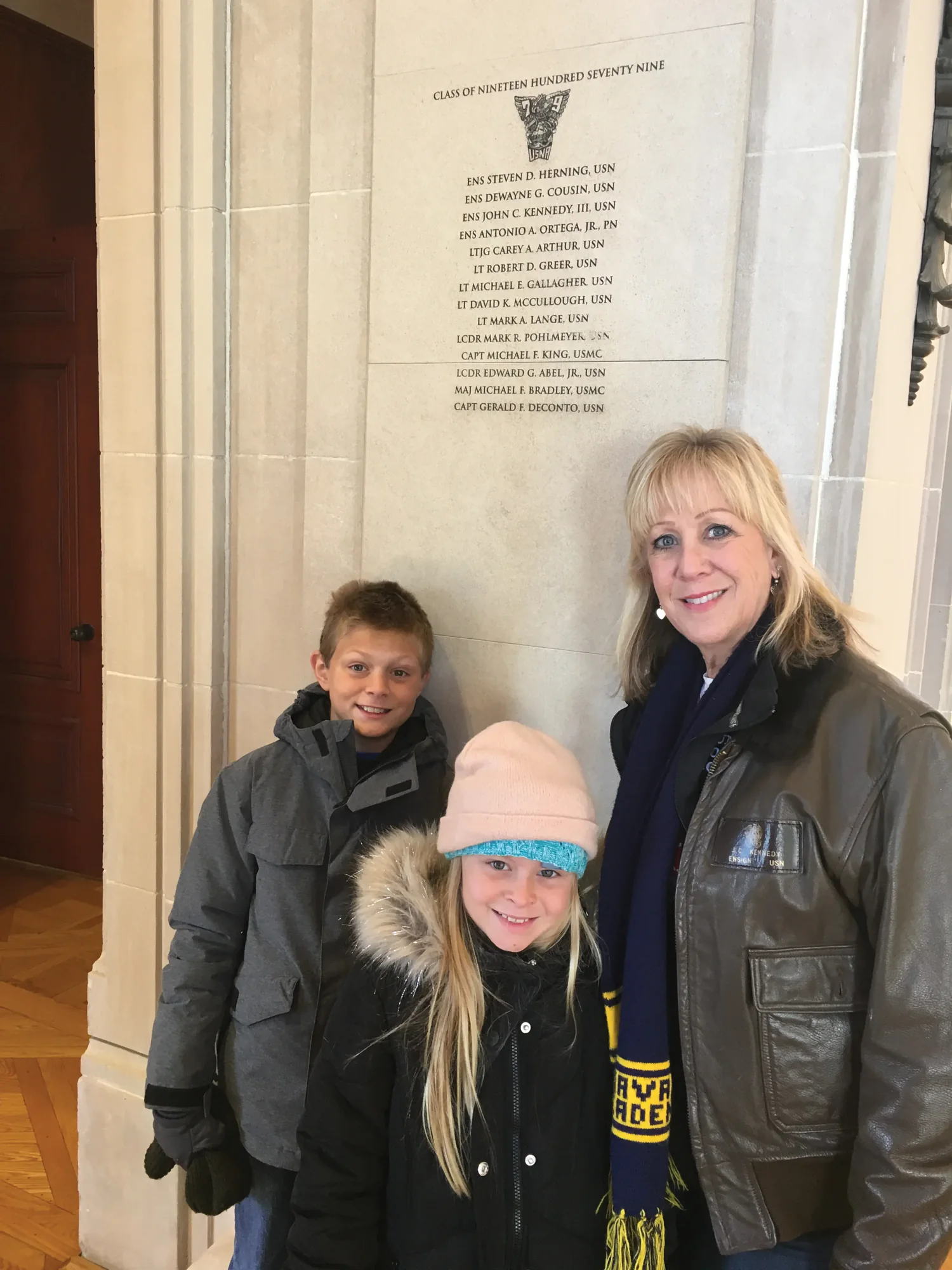 A woman in a flight jacket and two children beside the Class of 1979 panel in Memorial Hall.