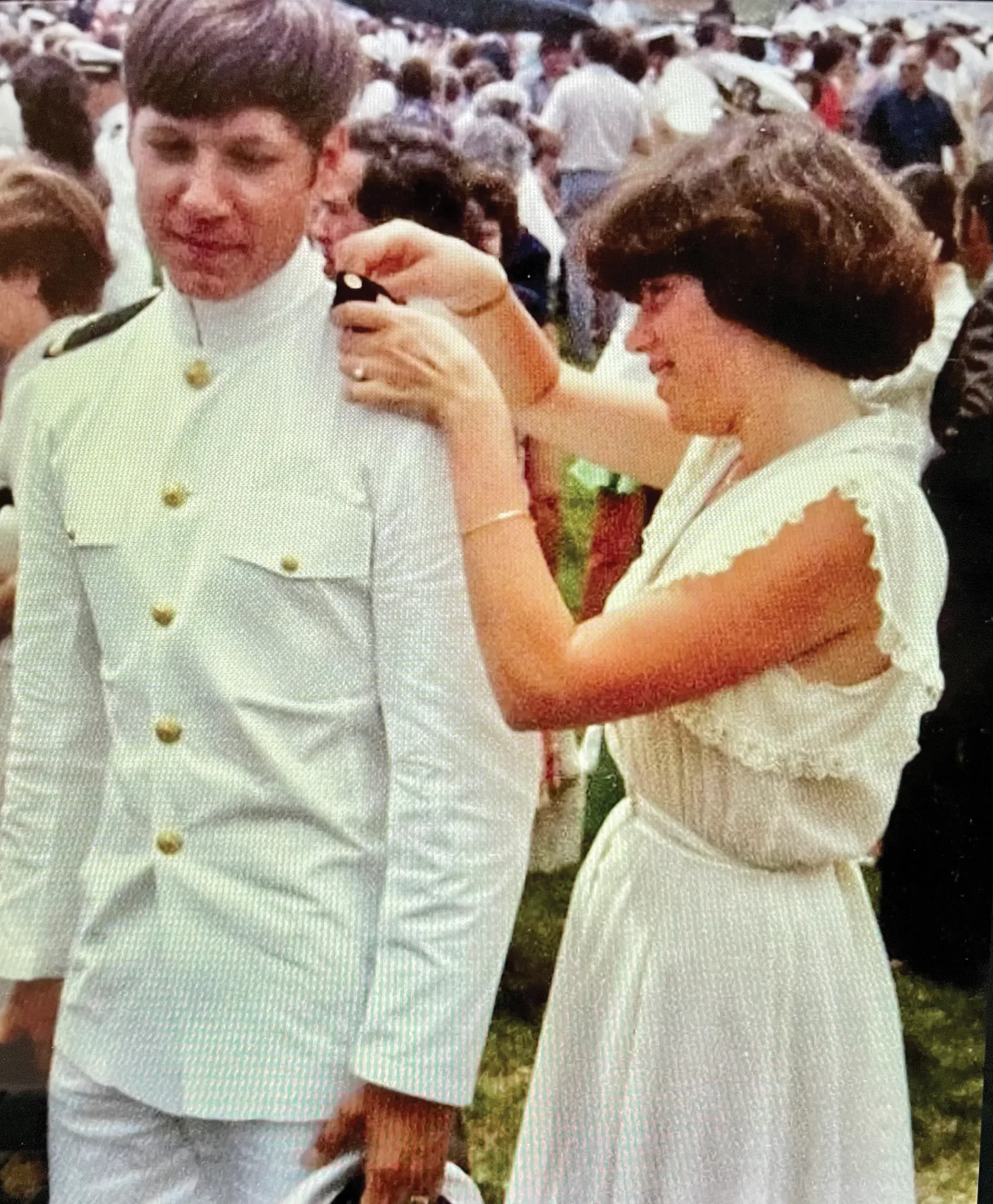 A young woman in a white dress pinning a shoulder board onto a midshipman in Summer Whites at a graduation ceremony.